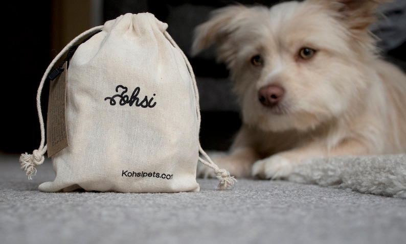 May include: A white canvas drawstring bag with the word "Zohsi" embroidered in black. The bag has a brown tag attached to it and the text "Kohsipets.com" printed on the bottom. A white dog with brown eyes is looking at the bag.