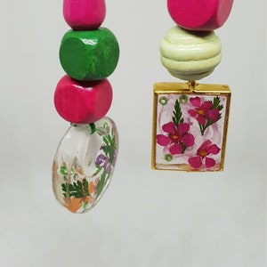 Puede incluir: Dos collares de cuentas coloridas con un esquema de colores verde y rosa. Un collar tiene un dije de resina transparente con flores prensadas. El otro collar tiene un dije de madera con un diseño floral.
