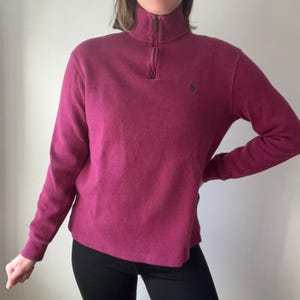 May include: A burgundy long-sleeved pullover sweater with a quarter-zip neckline and a small embroidered logo on the chest. The sweater has a ribbed collar and cuffs. The model is wearing black trousers.