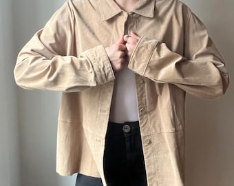 May include: A tan suede jacket with a collared neckline and button closures. The jacket has long sleeves and a relaxed fit. The person is wearing a white top and black pants. The jacket appears to be a vintage or retro style.
