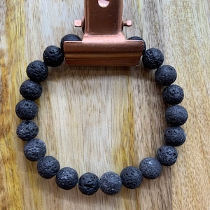 Op de afbeelding: Een zwarte lavasteen armband met een koperkleurige clip. De armband bestaat uit ronde, poreuze zwarte kralen. De clip is bevestigd aan de bovenkant van de armband, die op een houten oppervlak ligt.