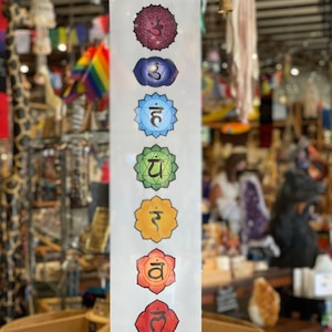 May include: A white banner featuring seven colorful chakra symbols, each with a unique design and color. The banner is displayed vertically and is held by wooden dowels at the top and bottom. The symbols are arranged from top to bottom in shades of purple, blue, green, yellow, orange, and red.