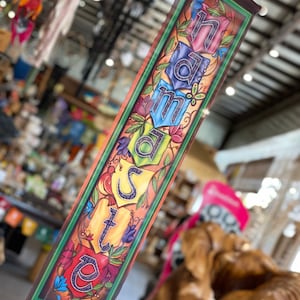 May include: A vertical banner with the word "Namaste" in colorful, stylized letters. The banner has a dark brown border with a green inner frame, and is decorated with floral and leaf motifs in shades of red, blue, and yellow.