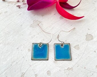 Puede incluir: Un par de pendientes de plata con colgantes cuadrados de esmalte azul. Los pendientes están sobre una superficie blanca con una flor rosa.