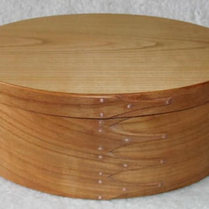 Può includere: Una scatola ovale in legno con una finitura in legno naturale. La scatola è realizzata in legno di ciliegio e ha un design semplice con accenti in rame.