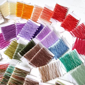 May include: Assortment of embroidery floss spools in various colors, including purple, green, brown, blue, red, and white. The spools are arranged in rows on a white surface.