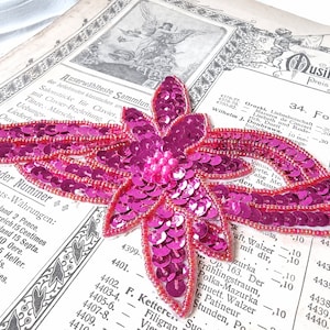 Può includere: Un'applicazione di fiori di paillettes rosa con un centro di perla. L'applicazione è su una pagina di un libro con testo in tedesco.