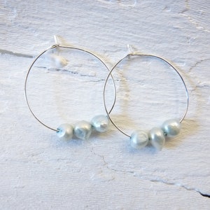 Può includere: Un paio di orecchini a cerchio d'argento con tre perle blu chiaro su ogni cerchio.