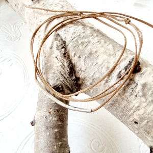 Può includere: Una semplice collana a barra d'argento con un cordoncino marrone. La collana è esposta su un pezzo di legno di deriva.