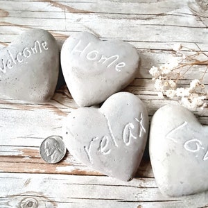 Pode incluir: Quatro pedras de concreto cinzas em forma de coração com as palavras "Welcome", "Home", "Relax" e "Love" inscritas nelas. As pedras estão dispostas sobre uma superfície de madeira clara.