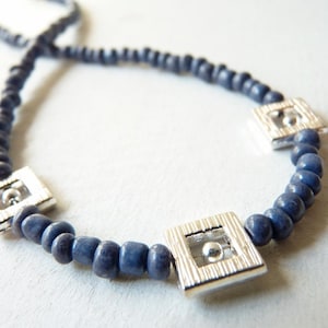Può includere: Una collana realizzata con perline blu scuro con accenti quadrati argentati. Ogni quadrato ha un piccolo cerchio al centro.
