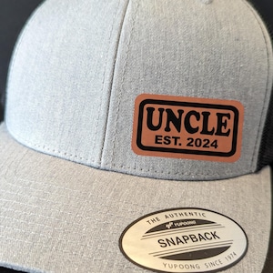 Può includere: Un cappello da baseball grigio e nero con una toppa in pelle marrone che dice "Uncle Est. 2024". Il cappello ha una parte posteriore in rete nera e una parte anteriore grigia con un logo bianco cucito che dice "The Authentic Yupoong Snapback Yupoong Since 1974".