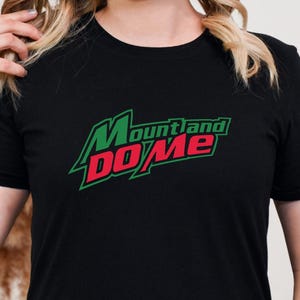 Pode incluir: T-shirt preto com o texto "Mount and Do Me" em verde e vermelho. A camisa tem gola redonda e mangas curtas. O texto está em uma fonte estilizada.