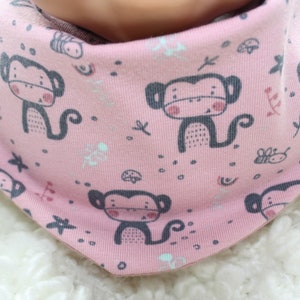 Könnte beinhalten: Rosa Baby-Lätzchen mit Affen-Motiv. Das Lätzchen hat ein kleines Affen-Design mit grauer Umrandung und einem rosa Gesicht. Das Lätzchen ist aus weichem Baumwollstoff gefertigt und perfekt, um die Kleidung des Babys sauber zu halten.