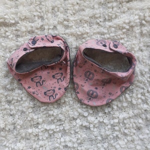 Könnte beinhalten: Zwei rosa Baby-Lätzchen mit einem Affen- und Sternmuster. Die Lätzchen haben ein graues Futter und einen Druckknopfverschluss. Sie sind aus weichem Stoff gefertigt und fangen Speichel und Verschüttetes auf.