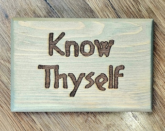 Puede incluir: Un letrero rectangular de madera con las palabras "Know Thyself" grabadas. El letrero tiene un lavado azul grisáceo claro sobre la madera, y el texto es de color marrón oscuro. El letrero se muestra sobre una superficie de madera.