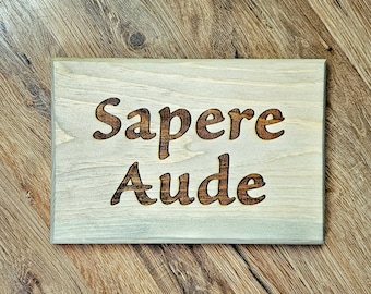 Puede incluir: Un letrero de madera con la frase latina "Sapere Aude" quemada en la superficie. El letrero es de color marrón claro con un acabado blanqueado.