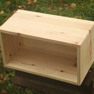 Könnte beinhalten: Eine rechteckige Holzkiste mit offener Vorderseite, aus hellem Holz gefertigt. Die Kiste steht auf einer dunklen Oberfläche vor dem Hintergrund von grünem Gras und Herbstlaub. Die Kiste dient wahrscheinlich zur Aufbewahrung oder Präsentation.