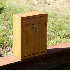 May include: A wooden mailbox with a keyhole and a key in the lock. The mailbox is made of light brown wood and has a simple design.