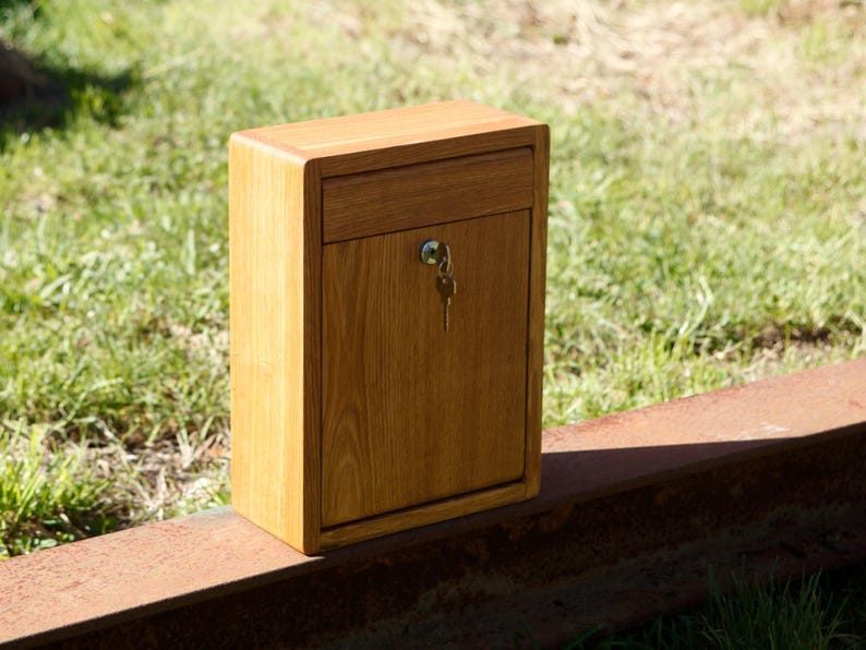May include: A wooden mailbox with a keyhole and a key in the lock. The mailbox is made of light brown wood and has a simple design.