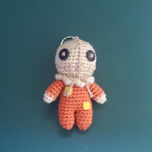 May include: A handmade crochet doll in the likeness of Sam from the horror film Trick 'r Treat. The doll features a beige head with button eyes, a stitched mouth, and an orange jumpsuit with yellow patches. The doll is approximately 4 inches tall.