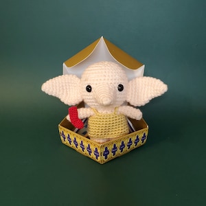May include: A crocheted pink and white house elf doll holding a red wand, sitting inside a golden box with blue and purple details.