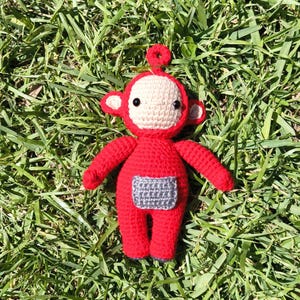 May include: A handmade crochet doll in the shape of a red Teletubby character. The doll has a beige face, black eyes, and a gray rectangle on its belly. The doll is lying on a bed of green grass.