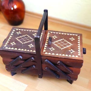 Könnte beinhalten: Eine Vintage-Nähkiste aus Holz mit dunkelbraunem Finish. Die Kiste hat ein dekoratives Intarsienmuster auf dem Deckel und ein Faltendesign. Der Griff ist dunkelbraun. Die Kiste wird wahrscheinlich zur Aufbewahrung von Nähutensilien verwendet.