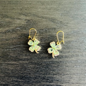 May include: A pair of gold-toned drop earrings featuring four-leaf clover charms. Each clover is crafted with light green translucent petals and accented with small clear stones along the edges, suspended from simple gold-toned ear wires.