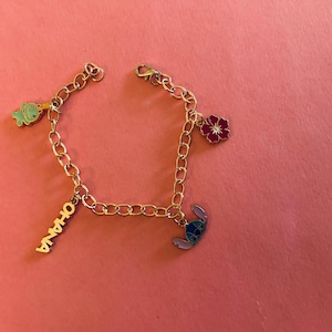 May include: A silver-colored charm bracelet with various enamel charms. Charms include a green dinosaur, a red flower, a blue alien, and a charm that says "OHANA". The bracelet is on a pink background.