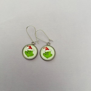 May include: A pair of silver-toned dangle earrings. Each earring features a circular charm with a green cartoon character wearing a red Santa hat. The earrings have a festive, holiday theme.