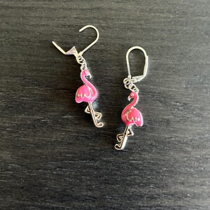 May include: A pair of silver-toned leverback earrings featuring dangling pink enamel flamingo charms. Each earring displays a small, bright pink flamingo with a curved neck and long legs, suspended from a secure metal leverback closure mechanism.