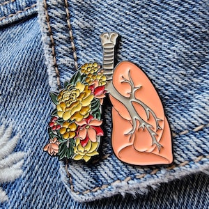 May include: A pink anatomical lung with a floral design. The lung is made of metal and has a silver-colored bronchial tree. The flowers are yellow, red, and green.