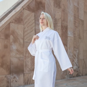 May include: A white bathrobe with long sleeves and a belt. The robe has a pocket on the left side and the initials "Y.S.K." embroidered on the chest.