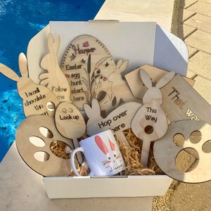 May include: A white box filled with wooden Easter egg hunt signs. The signs feature bunny rabbits, paw prints, and phrases like "I smell chocolate this way", "Follow me", "Look up", "Hop over here", "This way", and "That way". The box also includes a white mug with a bunny rabbit design and the name "Harper" on it.