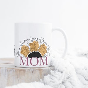 May include: White ceramic mug with a sunflower design and the word "MOM" in pink lettering. The sunflower has yellow petals and a black center. The words "Fearless", "Loving", "Warm", "Strong", and "Selfless" are written around the sunflower.