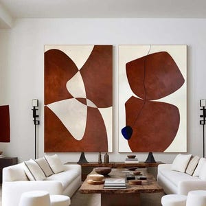 May include: A modern living room featuring two large abstract paintings with brown and white shapes. The room includes white sofas, a wooden coffee table, and white ottomans. The overall aesthetic is minimalist and contemporary.