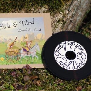 May include: A square cardboard CD case with a light brown finish and a printed illustration of two figures with a donkey and cart, featuring the text "Erde & Wind" and "Durch das Land". Beside it lies a black CD with white handwritten text.