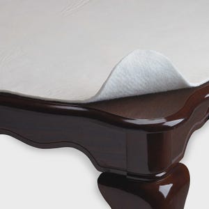 May include: A close-up of a dark brown wooden table with a white felt table protector. The table has a glossy finish and a curved edge. The felt protector is partially lifted, revealing the table's surface.