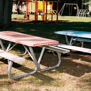 May include: Two picnic tables with red and white checkered tablecloths and blue and white checkered tablecloths on a grassy lawn.