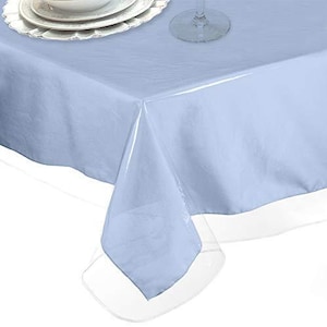 May include: A pale blue rectangular tablecloth with a clear vinyl border. The tablecloth is set on a table with white plates and a wine glass visible in the background. The tablecloth is made of a soft, smooth material.