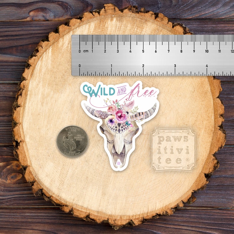 May include: A white skull with a floral crown and the words "WILD AND free" in a teal blue and pink font. The skull is on a wood background with a coin and a small sign that says "paws itivi tee".