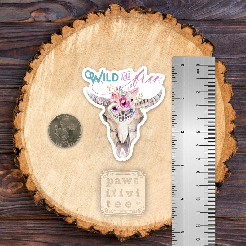 May include: A sticker featuring a watercolor illustration of a cow skull with a floral crown. The skull is white with black details and the flowers are pink, purple, and blue. The text "WILD AND free" is written in a whimsical font above the skull. The sticker is on a wooden background.