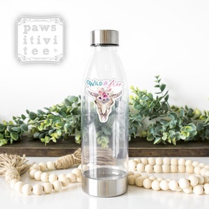 May include: A clear plastic water bottle with a stainless steel lid and base. The bottle has a sticker on it that says "Wild and Free" with a floral skull design.