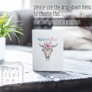 May include: A white notecard with a watercolor illustration of a cow skull with flowers and the text "WILD and Free". The notecard is sitting on a wooden table.
