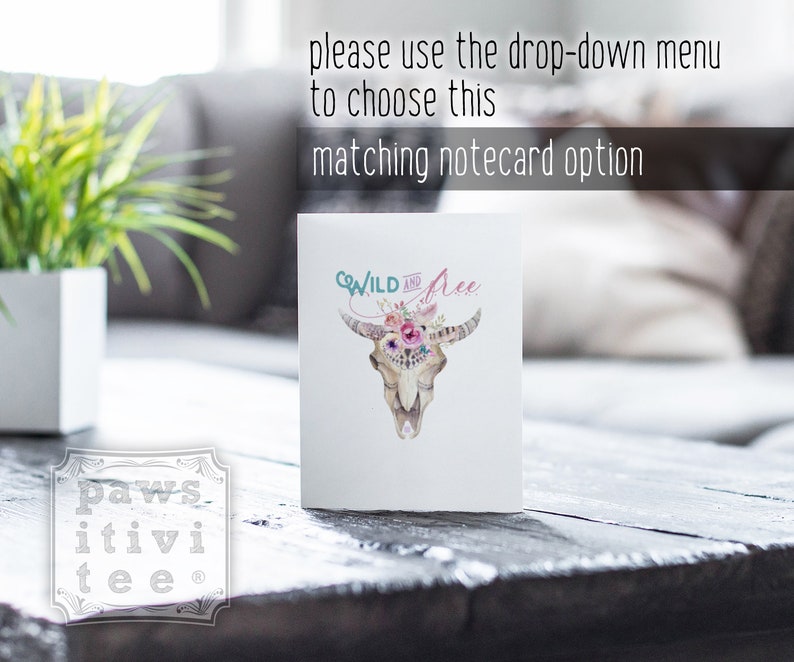 May include: A white notecard with a watercolor illustration of a cow skull with flowers and the text "WILD and Free". The notecard is sitting on a wooden table.