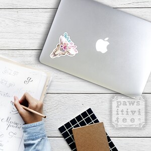 May include: A white laptop with a sticker of a skull with flowers and the text "Wild and Free". The laptop is on a wooden surface with a notebook and a pen.