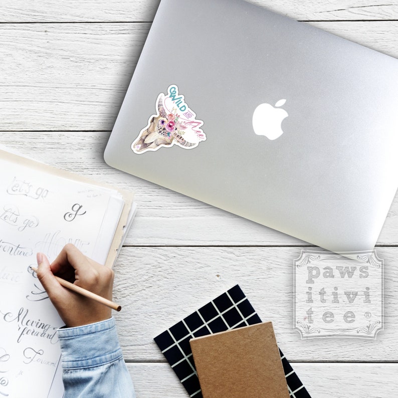 May include: A white laptop with a sticker of a skull with flowers and the text "Wild and Free". The laptop is on a wooden surface with a notebook and a pen.