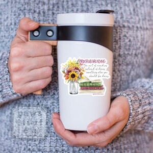 Pu&ograve; includere: Una tazza da viaggio bianca con coperchio nero e manico in legno. La tazza presenta una grafica con un bouquet di girasoli e foglie autunnali in un barattolo di vetro accanto a una pila di libri. Il testo recita: PROCRASTIREADING: the art of reading instead of doing something else you should be doing.