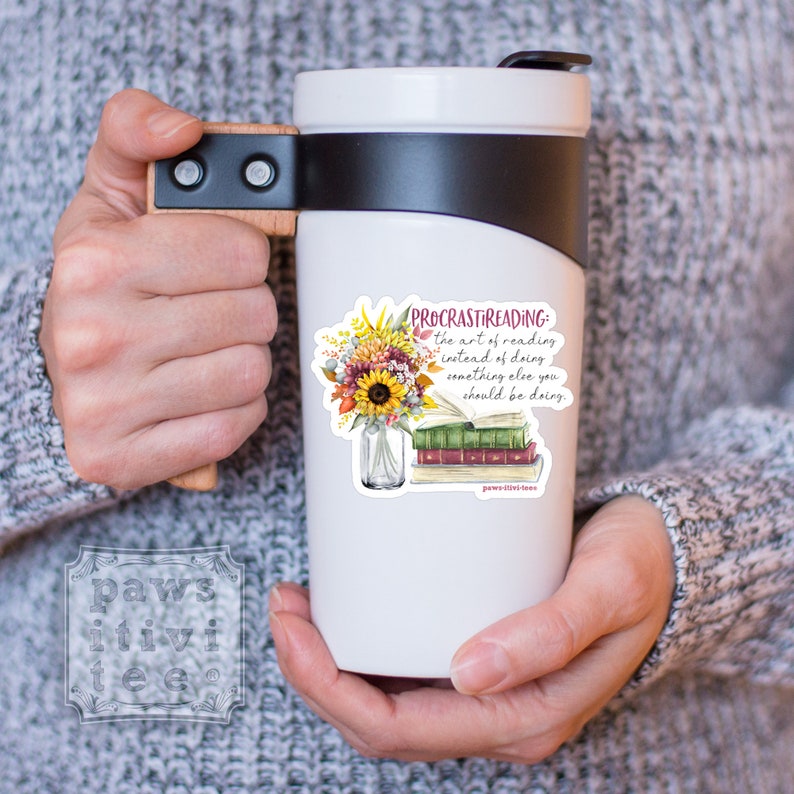 Pu&ograve; includere: Una tazza da viaggio bianca con coperchio nero e manico in legno. La tazza presenta una grafica con un bouquet di girasoli e foglie autunnali in un barattolo di vetro accanto a una pila di libri. Il testo recita: PROCRASTIREADING: the art of reading instead of doing something else you should be doing.